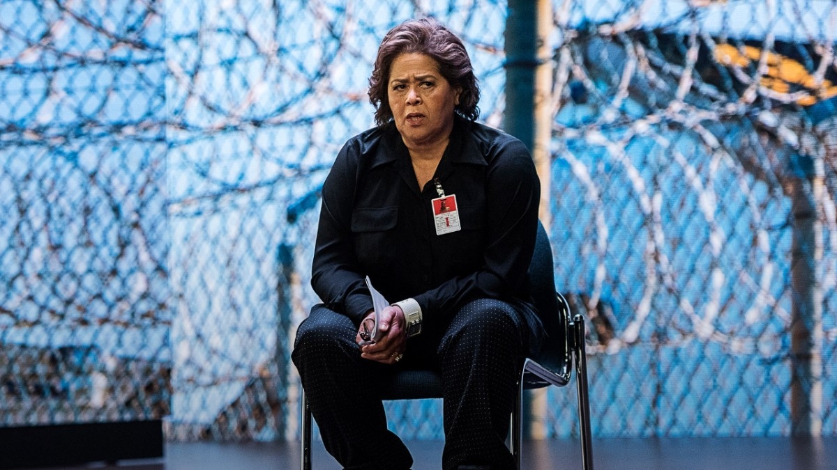 Anna Deavere Smith sits on chair in front of blue background with images of barbed wire